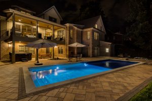 A well lit backyard at night with large tiled area and illuminated swimming pool with auto cover deploying
