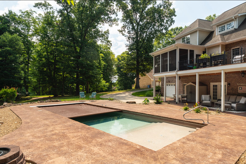 Rectangular inground pool with large tanning ledge and an automatic cover that is only partially retracted behind a large three story house with mature hardwoods