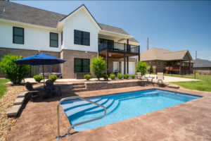 Very neatly landscaped pool area set outside a daylight basement with lounge chairs and small retaining wall