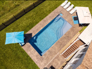 Overhead drone view of large fiberglass pool installation behind house and by the edge of a meadow with wildflowers