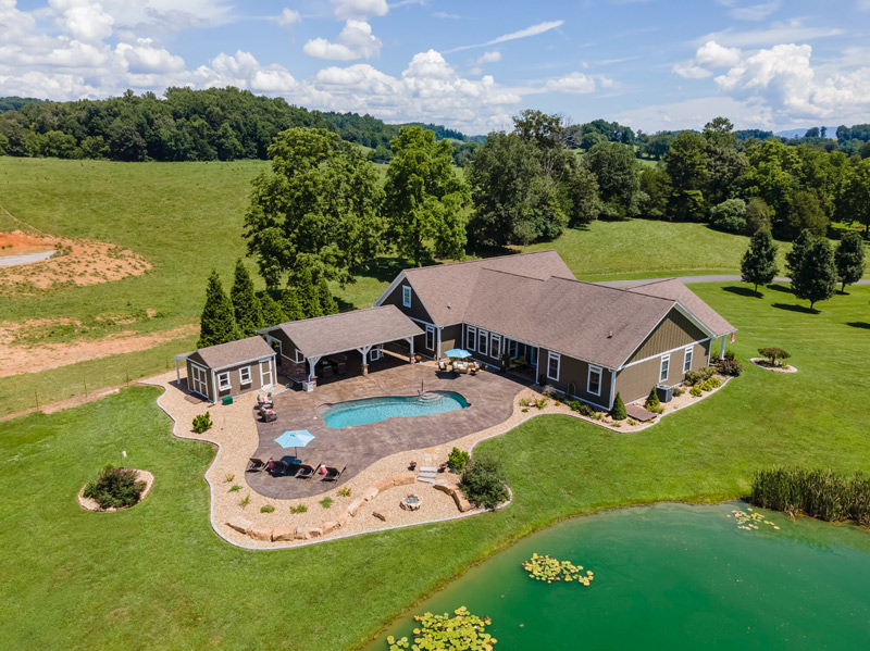 Aerial photography of swimming pool compound between house and pond, including covered outdoor living area, mechanicals shed, boulder wall, firepit area and expansive concrete decking