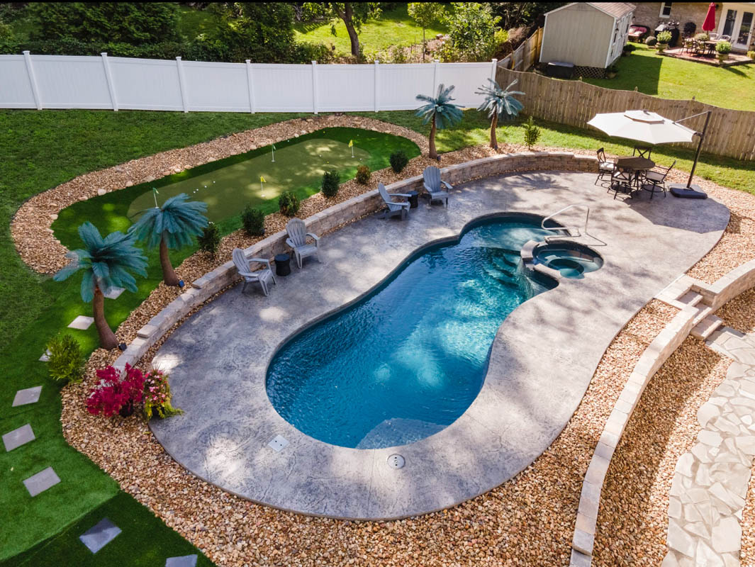 Overhead view of palm trees, inground pool, and putting green in backyard with tall white privacy fence