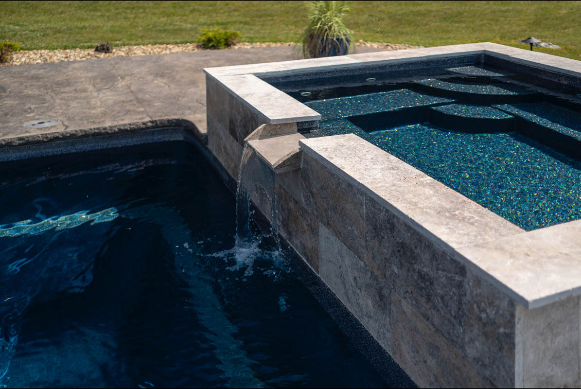 Jetted soaker tub skirted in silver travertine raised at pools edge with a spillover waterfall