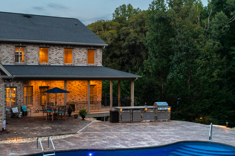 The outdoor kitchen, dining area, and back porch illuminated at dusk behind this Rogersville home